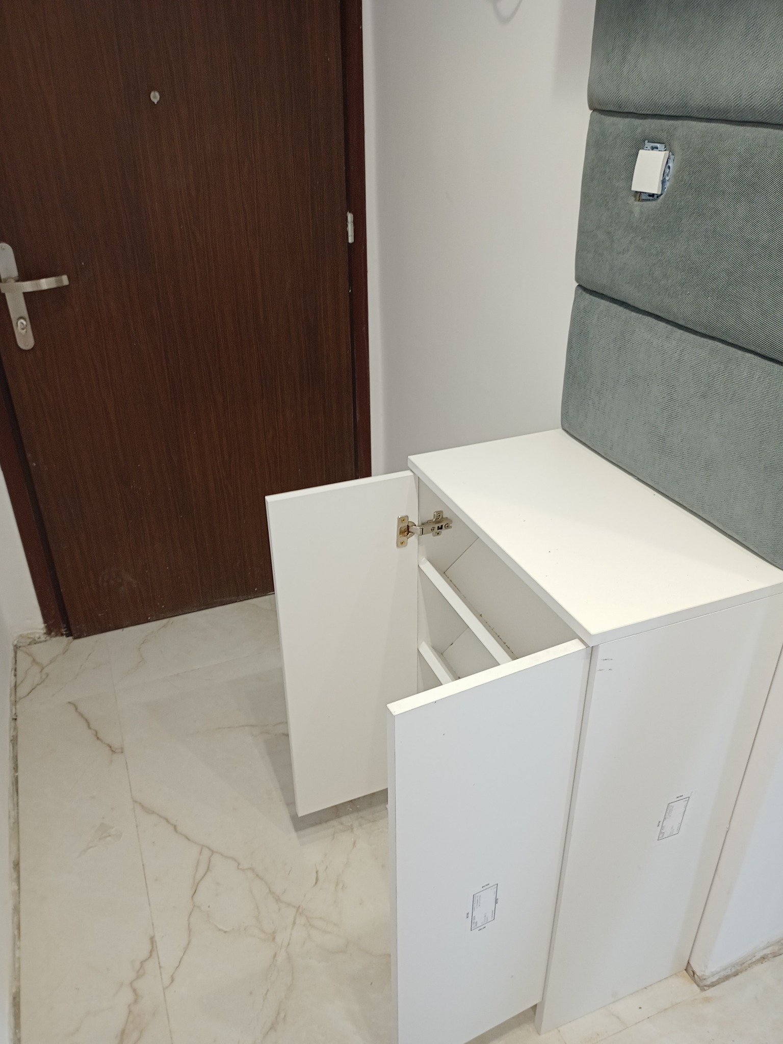 Biała szafka z otwartymi drzwiczkami, widoczne półki wewnątrz, obok brązowe drzwi i tapicerowany panel ścienny w kolorze szarym.