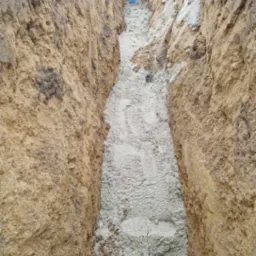 Wykop w ziemi z ułożoną niebieską rurą na dnie, zasypany piaskiem, widok z góry.