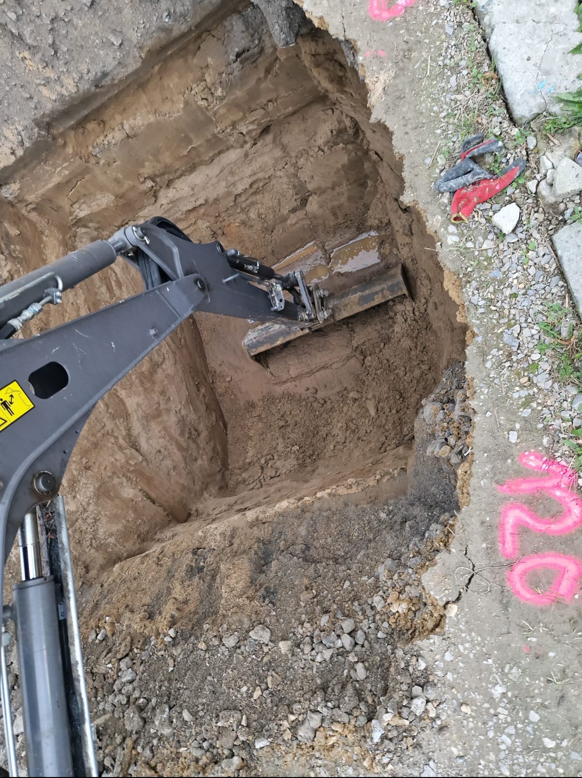 Koparka w trakcie wykonywania wykopu w ziemi, widoczne ślady po pracy łyżki, obok fragment chodnika z oznaczeniami w kolorze różowym.