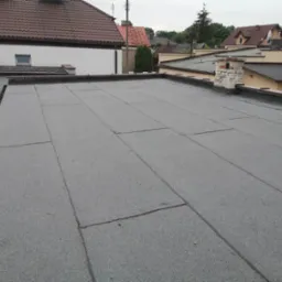Szary, płaski dach pokryty papą termozgrzewalną, widoczne łączenia papy, w tle budynki mieszkalne i uszkodzony komin z cegły.