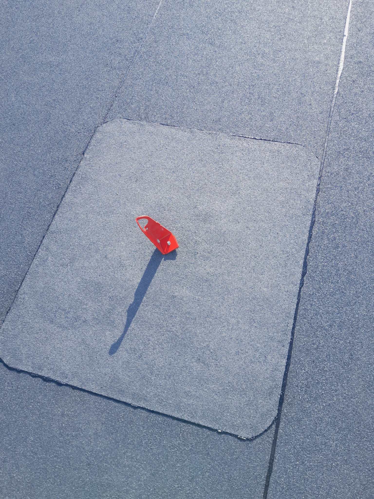 Fragment pokrycia dachowego z papy bitumicznej z widocznym włazem i czerwonym uchwytem do otwierania, rzucającym cień.