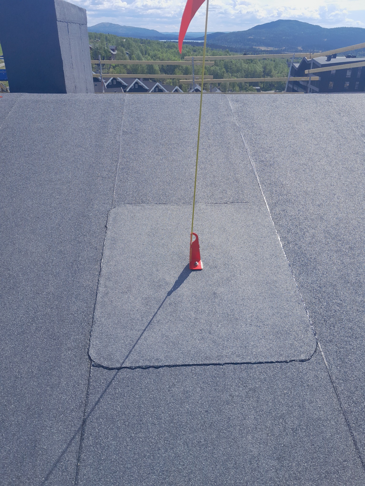 Nowe, płaskie pokrycie dachu z papy bitumicznej, widoczny fragment komina, zabezpieczenie krawędzi dachu i czerwony znacznik na żółtym sznurku.