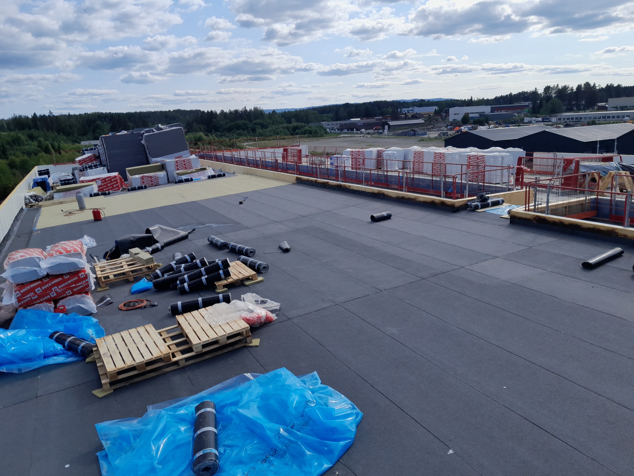 Rozległy płaski dach pokryty czarną papą termozgrzewalną, z widocznymi rolkami papy na paletach, materiałami izolacyjnymi w oddali i zabezpieczeniami krawędzi dachu, na tle zieleni i zabudowań.