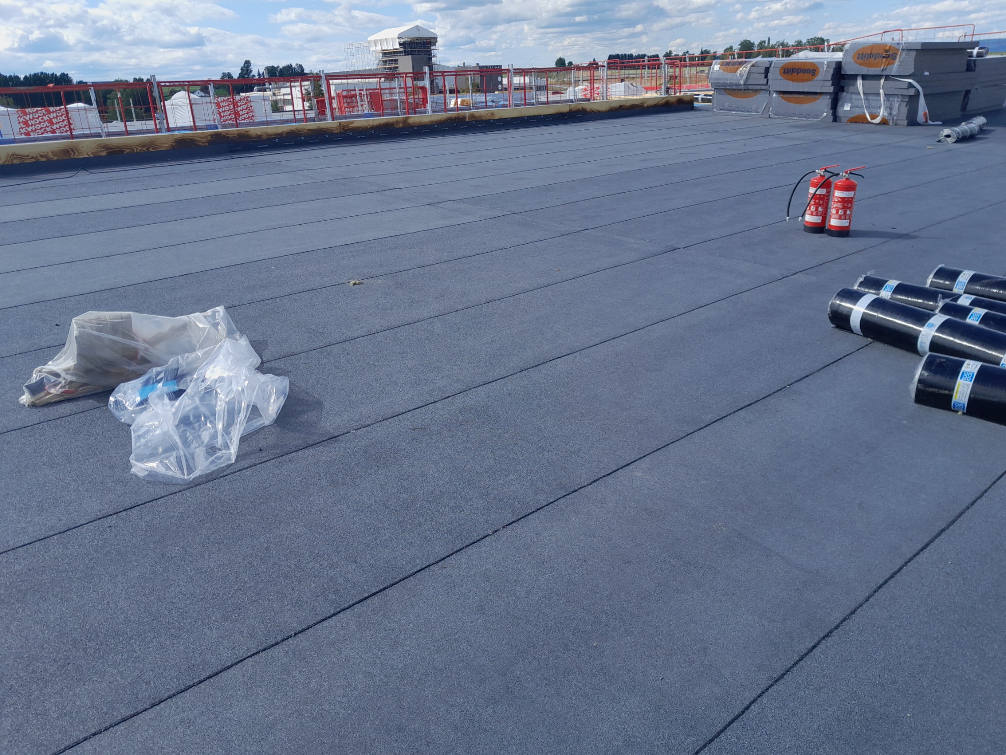 Nowa, płaska powierzchnia dachu pokryta papą termozgrzewalną, z widocznymi rolkami papy, dwoma gaśnicami i workiem foliowym. W tle zabezpieczenia na krawędzi dachu oraz materiały budowlane.