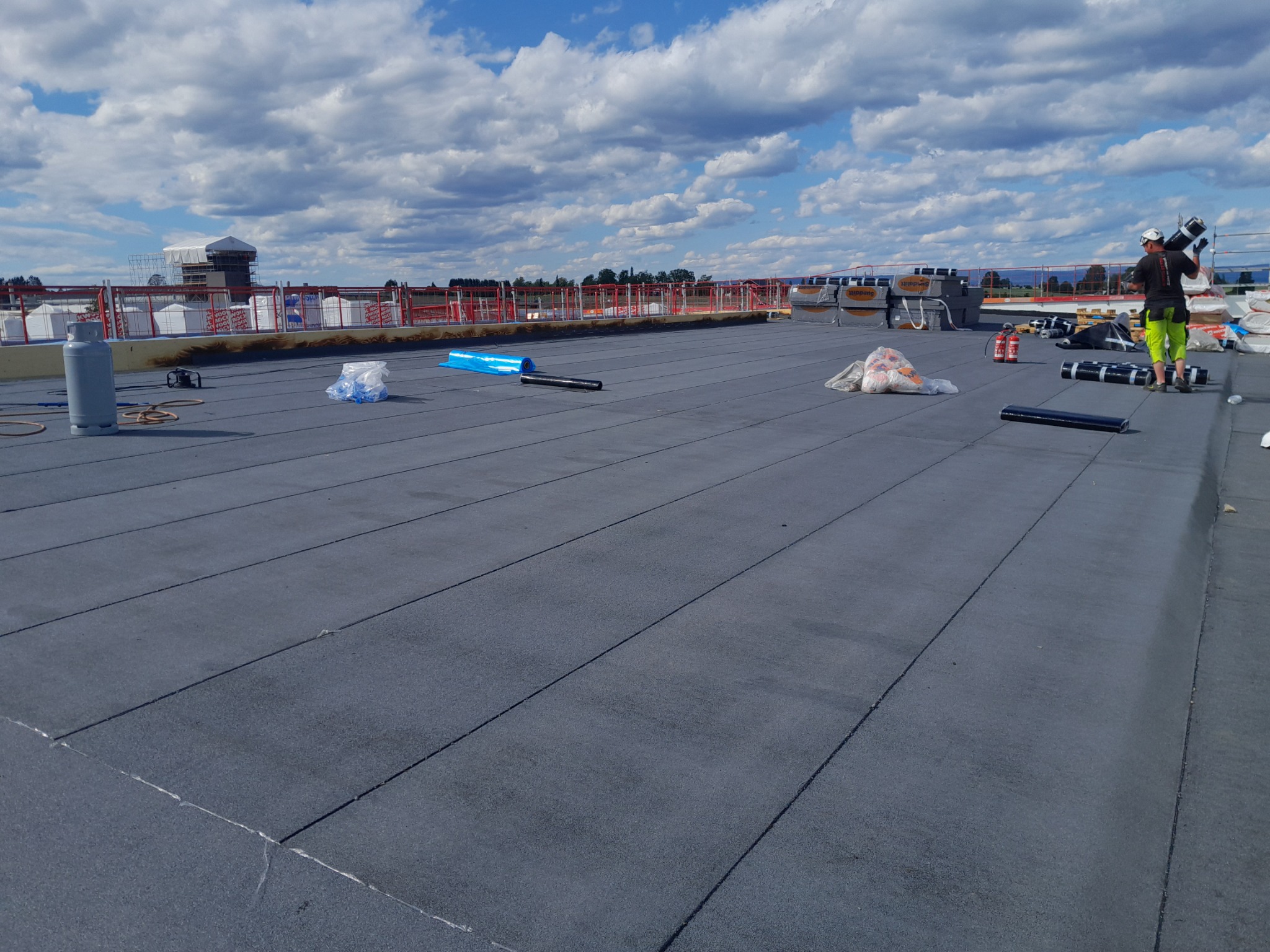 Czarny płaski dach pokryty papą termozgrzewalną z widocznymi zgrzewami, na dachu rozłożone materiały budowlane i butla gazowa, w tle pracownik w odblaskowej kamizelce niosący rolkę papy