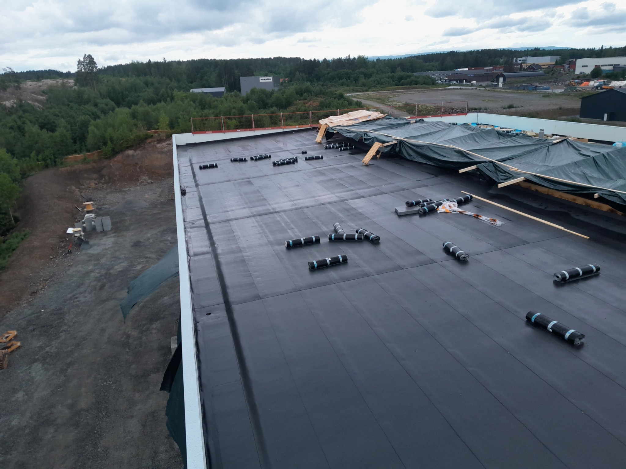 Czarna, płaska powierzchnia dachu pokryta papą, częściowo zabezpieczona plandeką, z widocznymi rolkami papy przygotowanymi do montażu i widokiem na okoliczne tereny zielone.