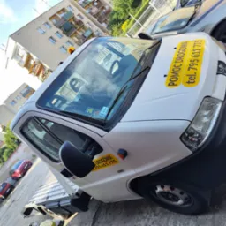 Biała laweta Peugeot z żółtymi naklejkami 'Pomoc Drogowa' i numerem telefonu na karoserii, zaparkowana na tle budynku mieszkalnego i innych samochodów.