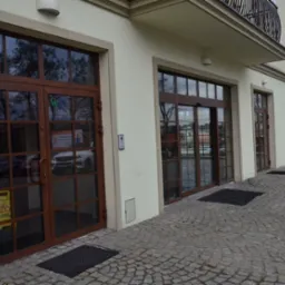 Elewacja budynku z trzema parami brązowych, przeszklonych drzwi z podziałami, ułożonych w rzędzie, z widoczną kostką brukową na chodniku i balkonem na piętrze.