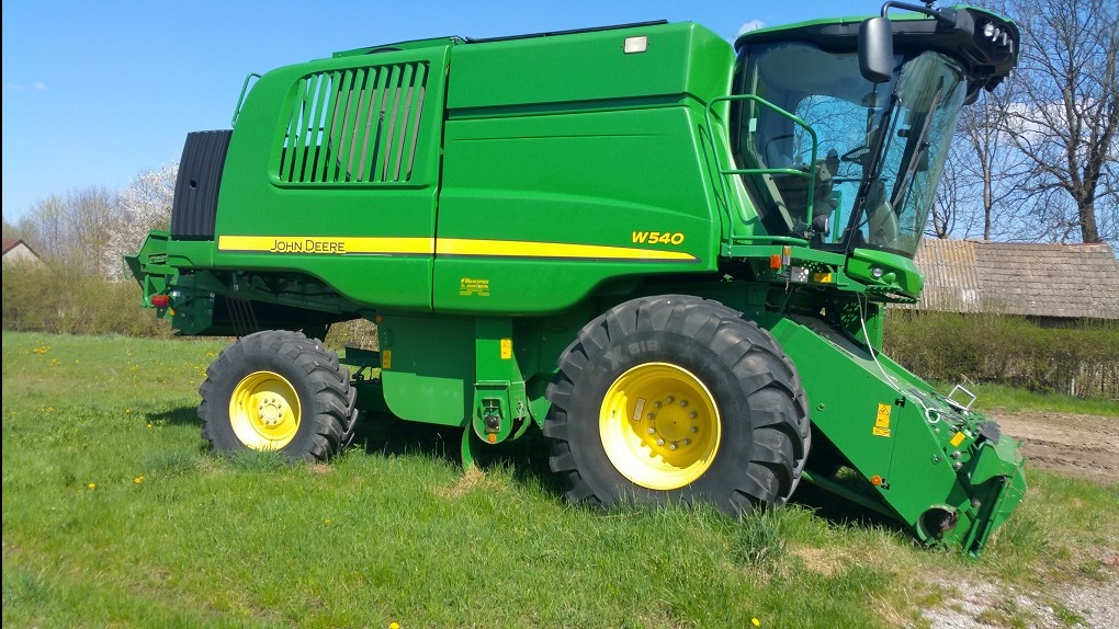 Zielony kombajn John Deere W540 na trawiastym polu, widok z boku, w tle budynek gospodarczy i drzewa.