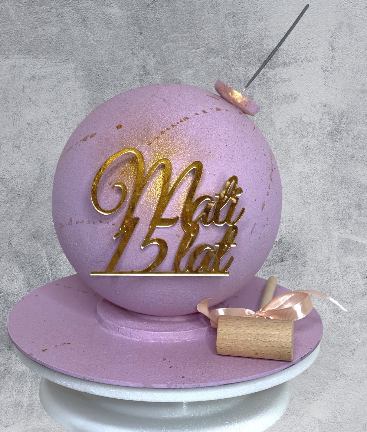Liliowy tort w kształcie kuli z napisem 'Mati 15 lat' wykonanym ze złotego akrylu, umieszczony na liliowej podstawie na białej paterze, obok mały drewniany młoteczek z różową wstążką.
