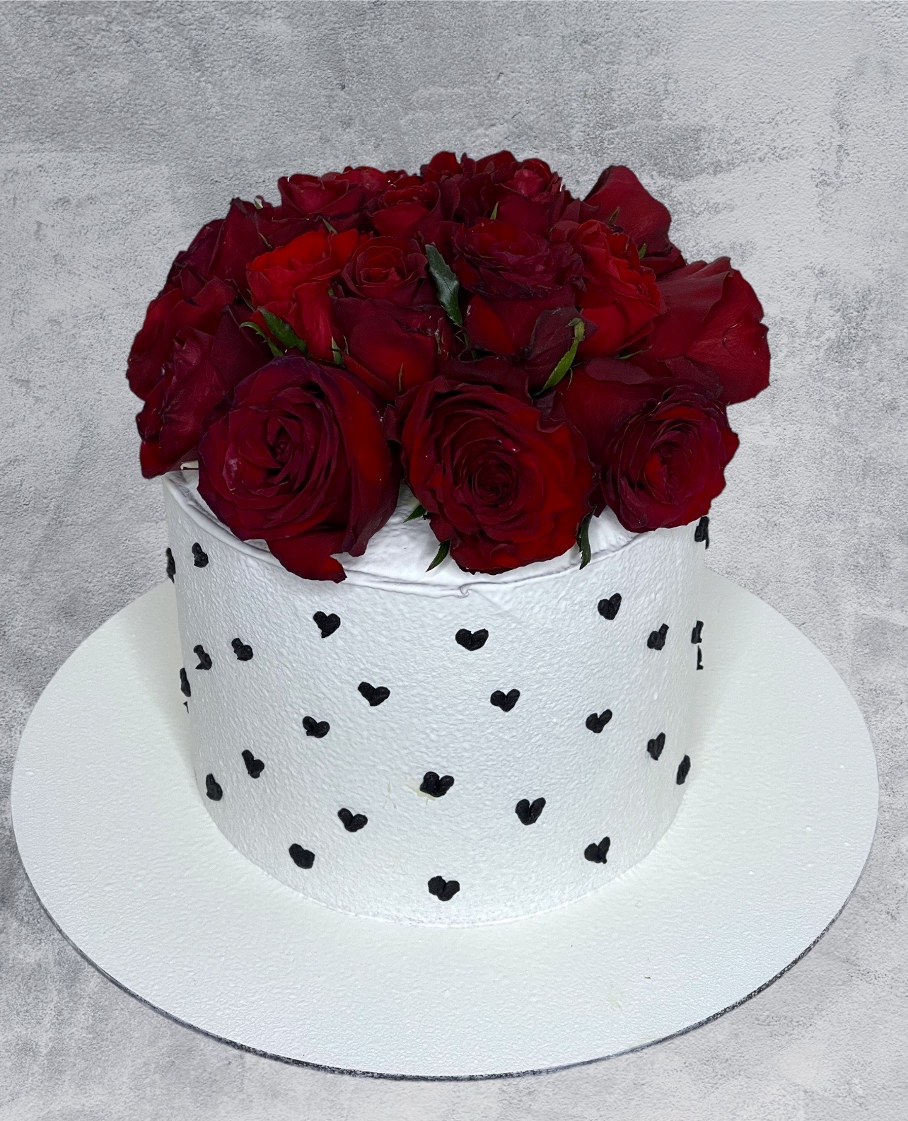 Tort z białym kremem zdobiony czarnymi serduszkami, udekorowany bukietem intensywnie czerwonych róż na wierzchu, umieszczony na białej paterze.