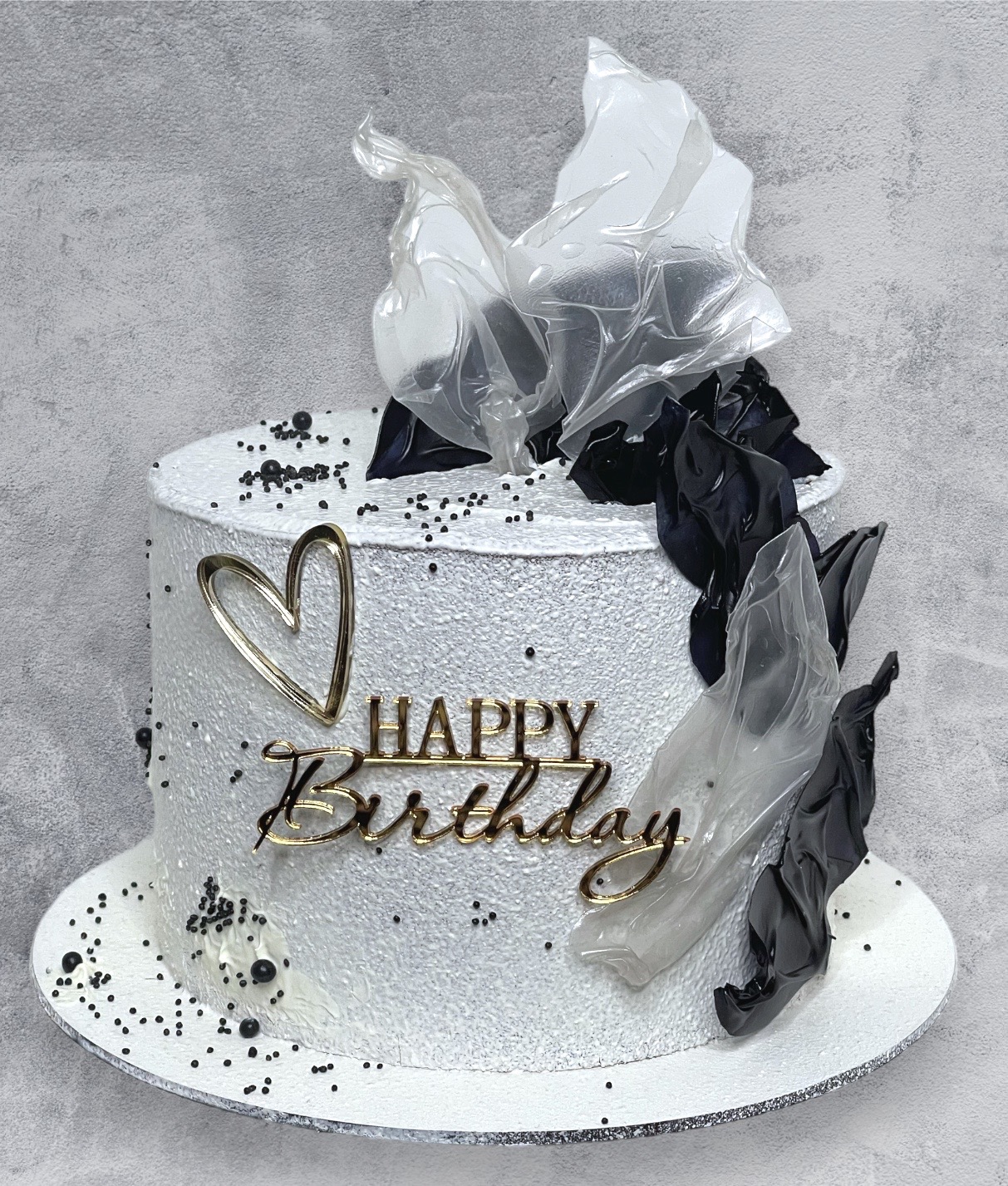 Tort urodzinowy z białym lukrem, złotym napisem 'Happy Birthday', sercem i dekoracjami z czarnej i przezroczystej folii, posypany czarnymi kuleczkami.
