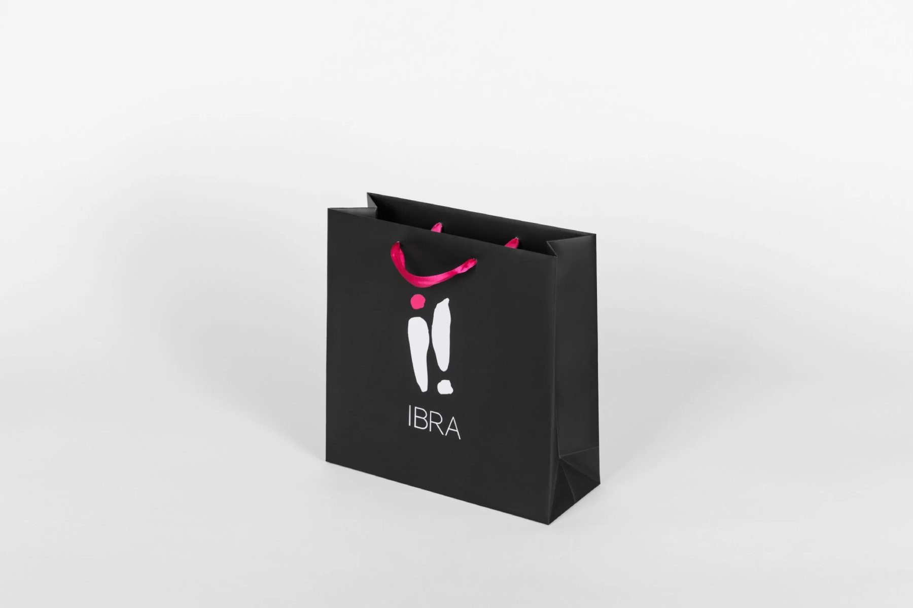 Czarna torba papierowa z białym logo i różowymi uchwytami sznurkowymi, ustawiona pod kątem na białym tle.