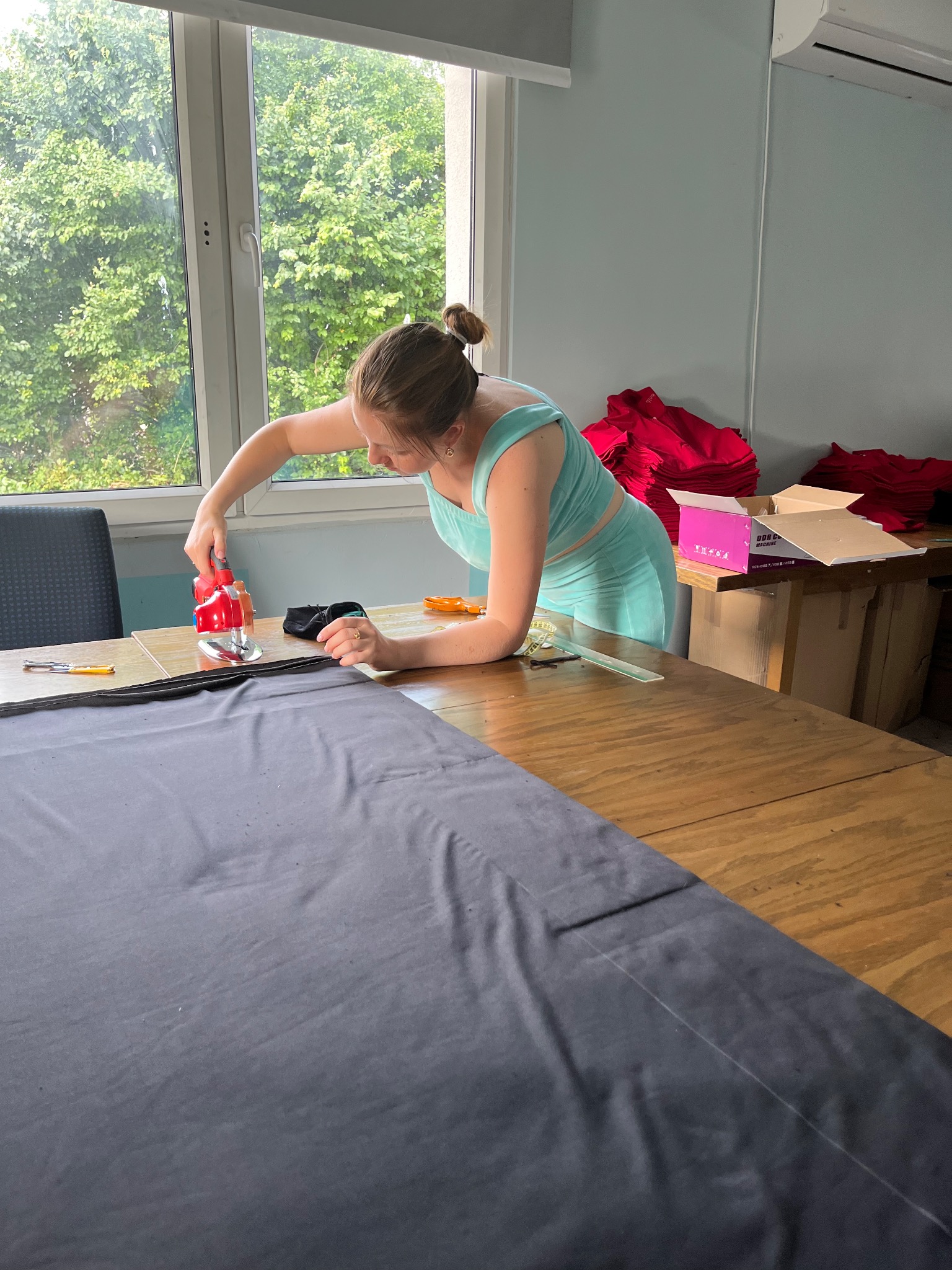 Kobieta w turkusowym stroju, stojąc pochylona nad dużym stołem, przycina szary materiał elektrycznym nożem krawieckim; w tle okno z widokiem na zieleń i stos czerwonych ubrań.