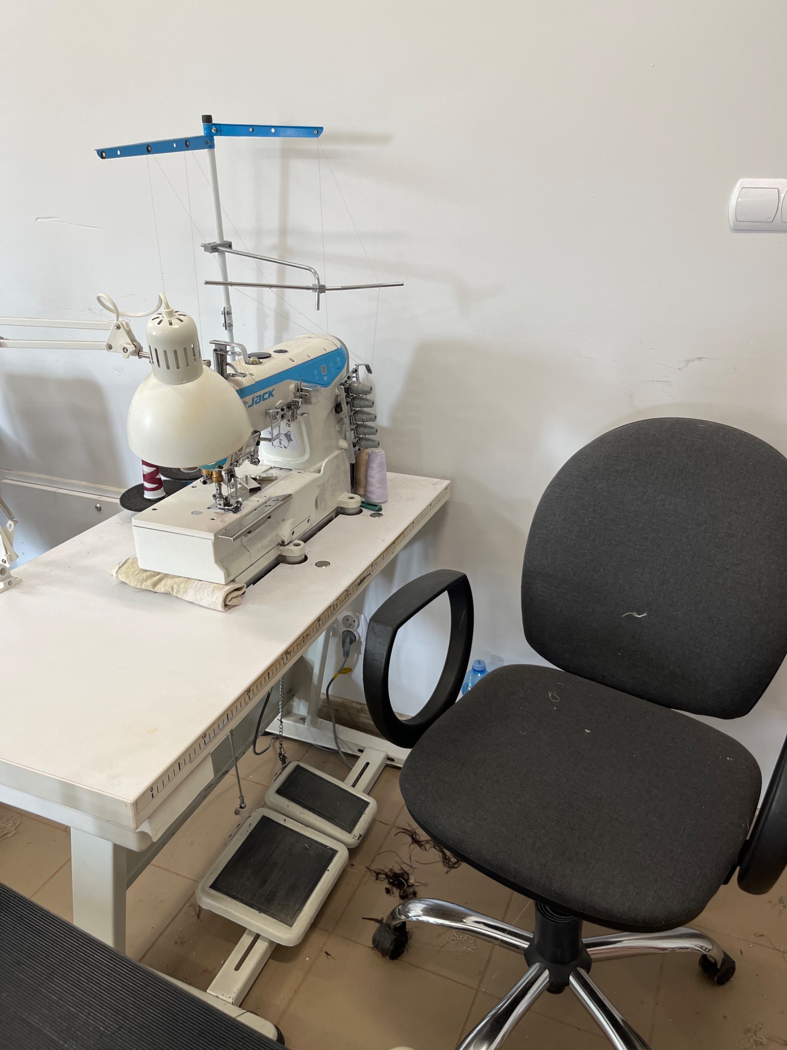 Stanowisko pracy w szwalni: maszyna do szycia Jack z lampą i pedałami na białym stole, obok krzesło biurowe, widoczne nitki i kurz na podłodze.