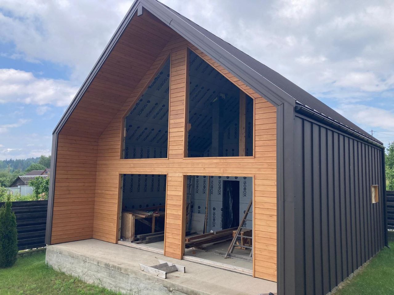 Budynek w trakcie budowy z drewnianą elewacją i czarnym metalowym dachem, widoczne okna z izolacją, częściowo wykończony, z elementami drewnianymi i metalowymi. Na podłodze widoczne materiały...