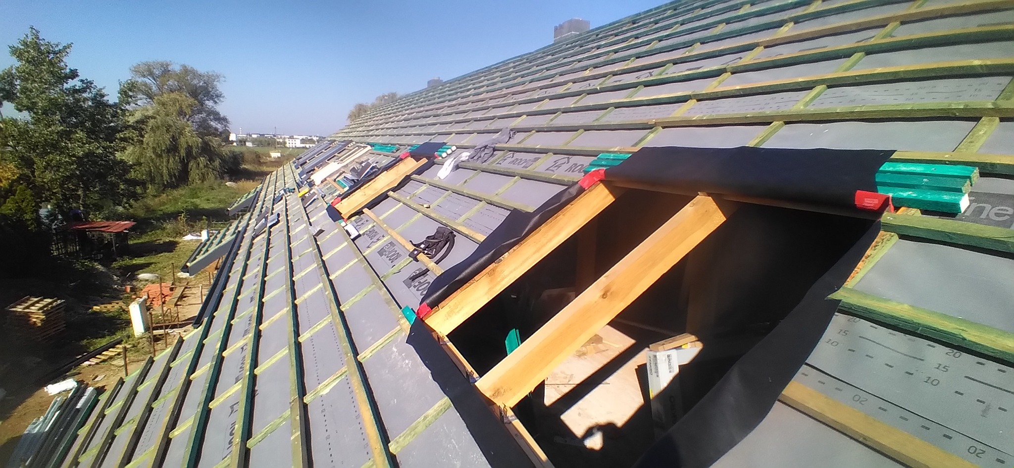 Widok z góry na konstrukcję dachu w trakcie budowy, z widocznymi łatami, membraną dachową z nadrukiem, elementami drewnianymi okien połaciowych i rolką czarnej folii w tle, na zewnątrz widoczne...