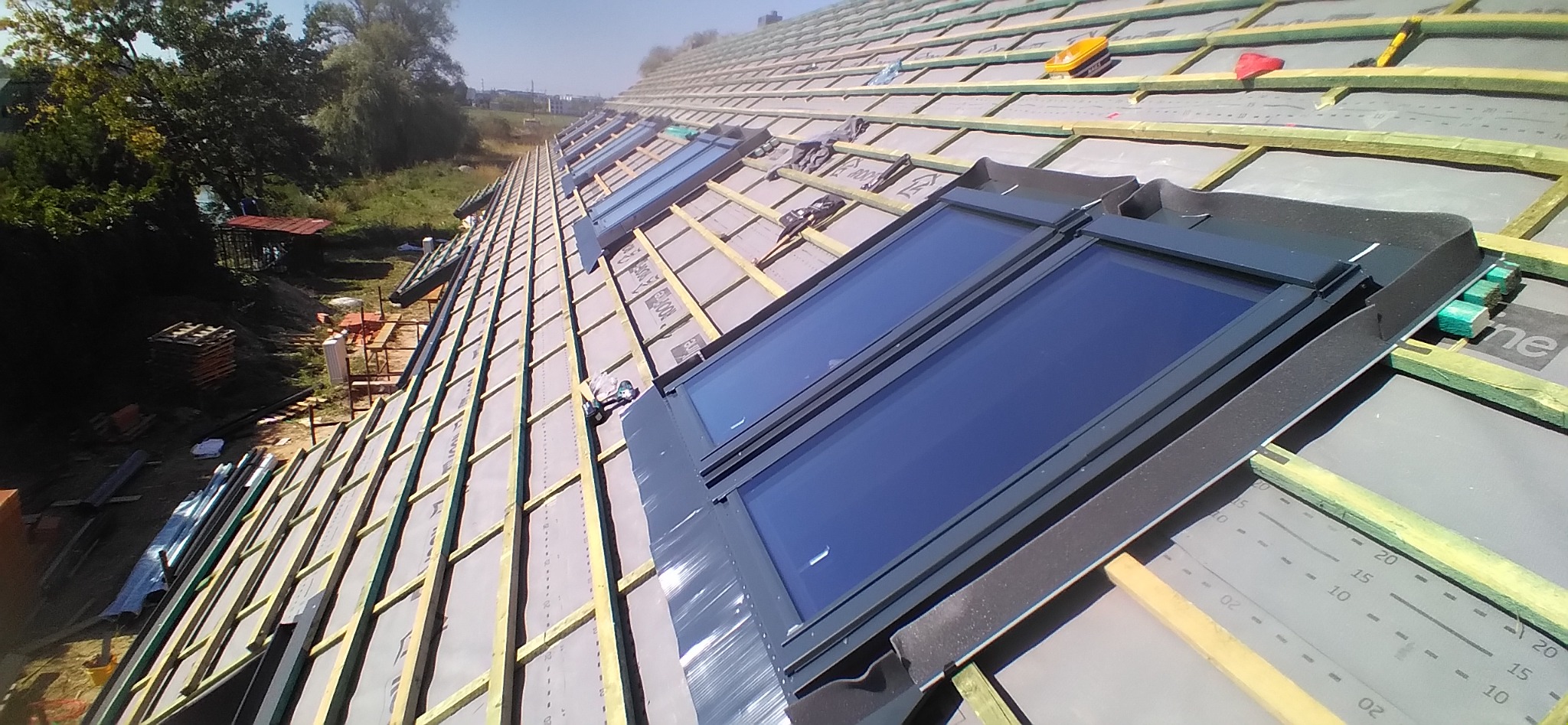 Montaż okien dachowych na nowym dachu z widoczną konstrukcją drewnianą i membraną dachową, w tle drzewa i zabudowania.