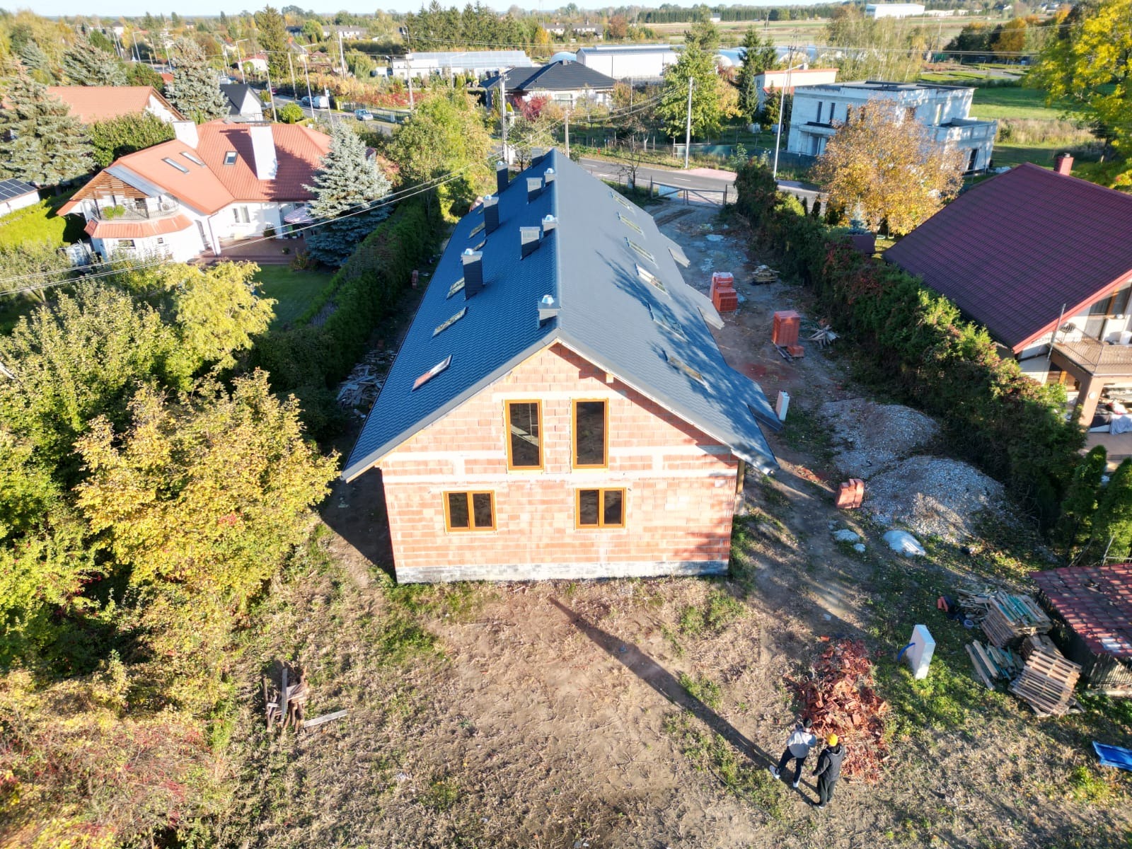 Widok z drona na budynek w trakcie budowy z ciemnoszarą dachówką, widoczne niezabudowane okna, mur z cegły, przygotowane materiały budowlane i dwie osoby w żółtym kasku na ziemi.