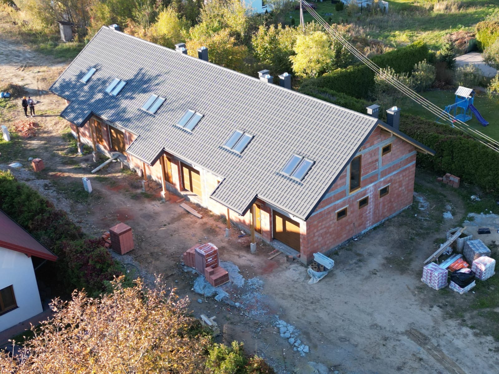 Wielorodzinny budynek w stanie surowym otwartym z szarym dachem z dachówką, widoczne okna dachowe i kominy, na zewnątrz porozrzucane materiały budowlane, ujęcie z drona.