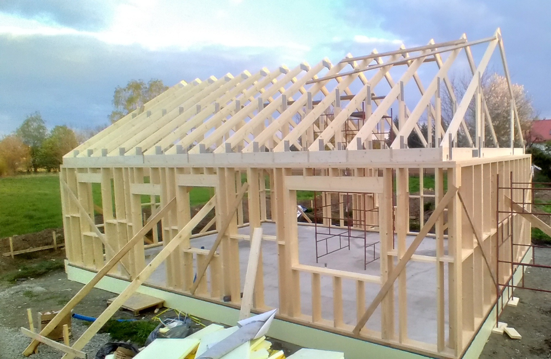 Surowa konstrukcja szkieletowa domu z drewna, widoczne elementy ścian, otwory okienne i dach, na zewnątrz zielony trawnik i drzewa.