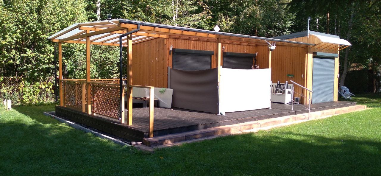 Drewniany domek letniskowy z zadaszonym tarasem, widoczne rolety zewnętrzne, ażurowa balustrada i rozkładane parawany na podeście, otoczony zielenią.