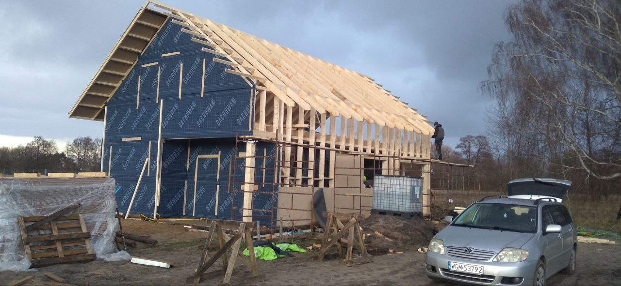 Wznoszenie szkieletu domu z drewnianych belek, ściany pokryte membraną dachową, widoczny pracownik na rusztowaniu, samochód osobowy z otwartym bagażnikiem na pierwszym planie.