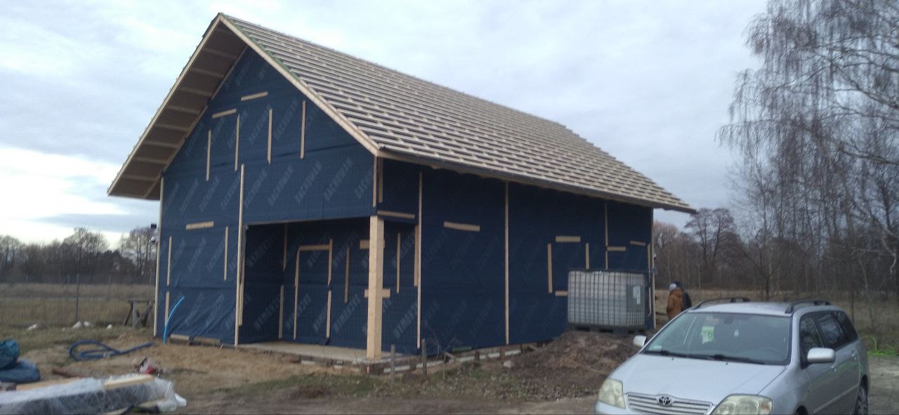 Budowa domu szkieletowego na etapie montażu łat dachowych i folii wiatroizolacyjnej na ścianach zewnętrznych, widoczny samochód osobowy i teren budowy.