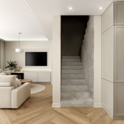 Inargo - Jasne wnętrze salonu z betonowymi schodami, beżową sofą i parkietem w jodełkę, minimalistyczny design, oświetlenie punktowe i wisząca lampa, szafki w zabudowie.