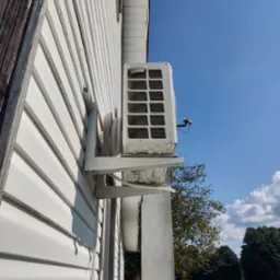 Zewnętrzna jednostka klimatyzatora zamontowana na białej ścianie domu z sidingu, widok z dołu na błękitne niebo, drabina oparta o ścianę w tle.