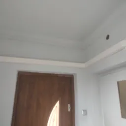 Wnętrze domu w trakcie remontu: widoczna drewniana ościeżnica drzwiowa z częściowo przeszklonymi drzwiami, biała listwa przypodłogowa pod sufitem i otwór w ścianie przygotowany pod instalację.