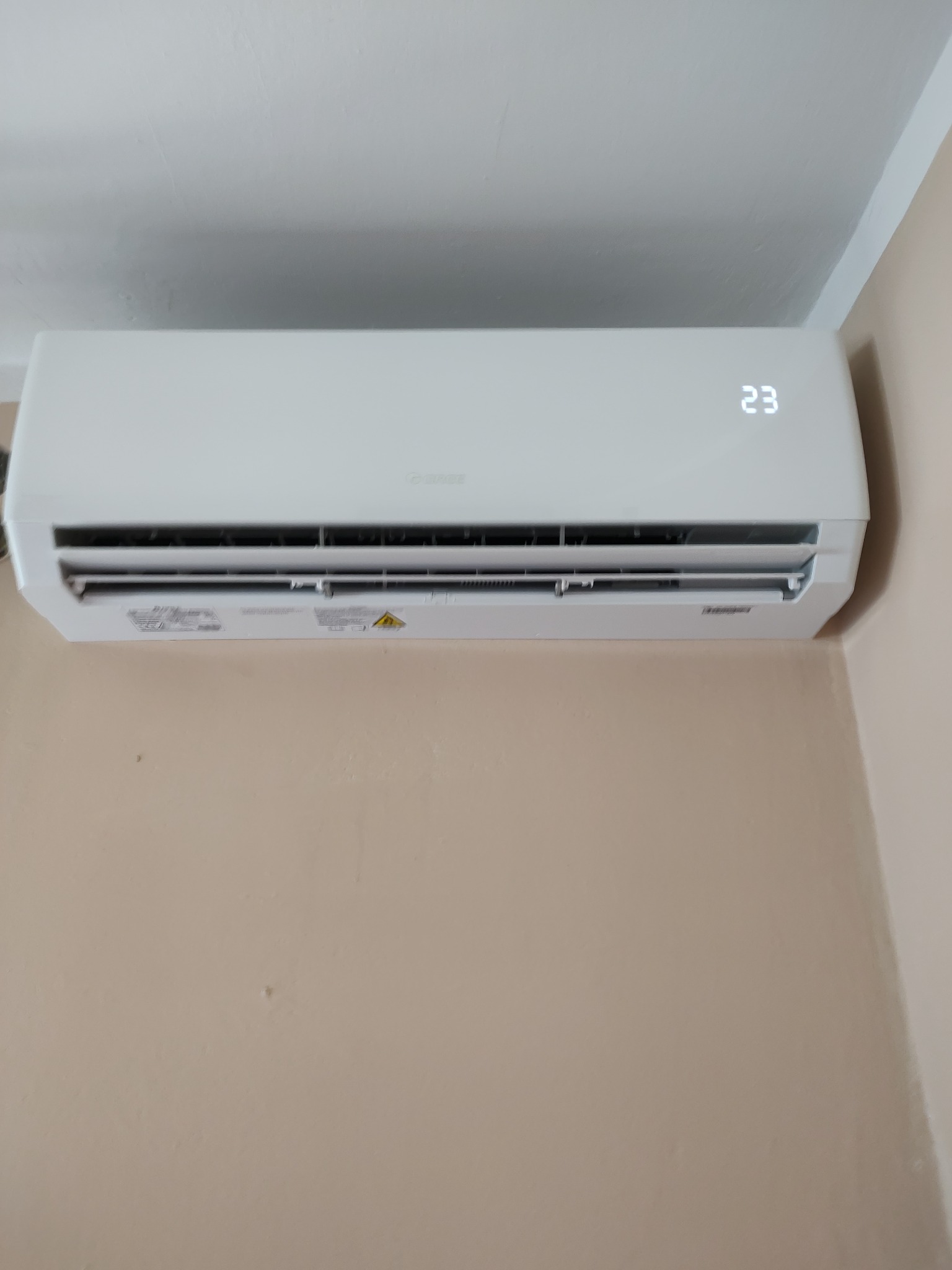 Biały klimatyzator ścienny z wyświetlaczem temperatury 23 stopnie Celsjusza, zamontowany na beżowej ścianie w rogu pomieszczenia.