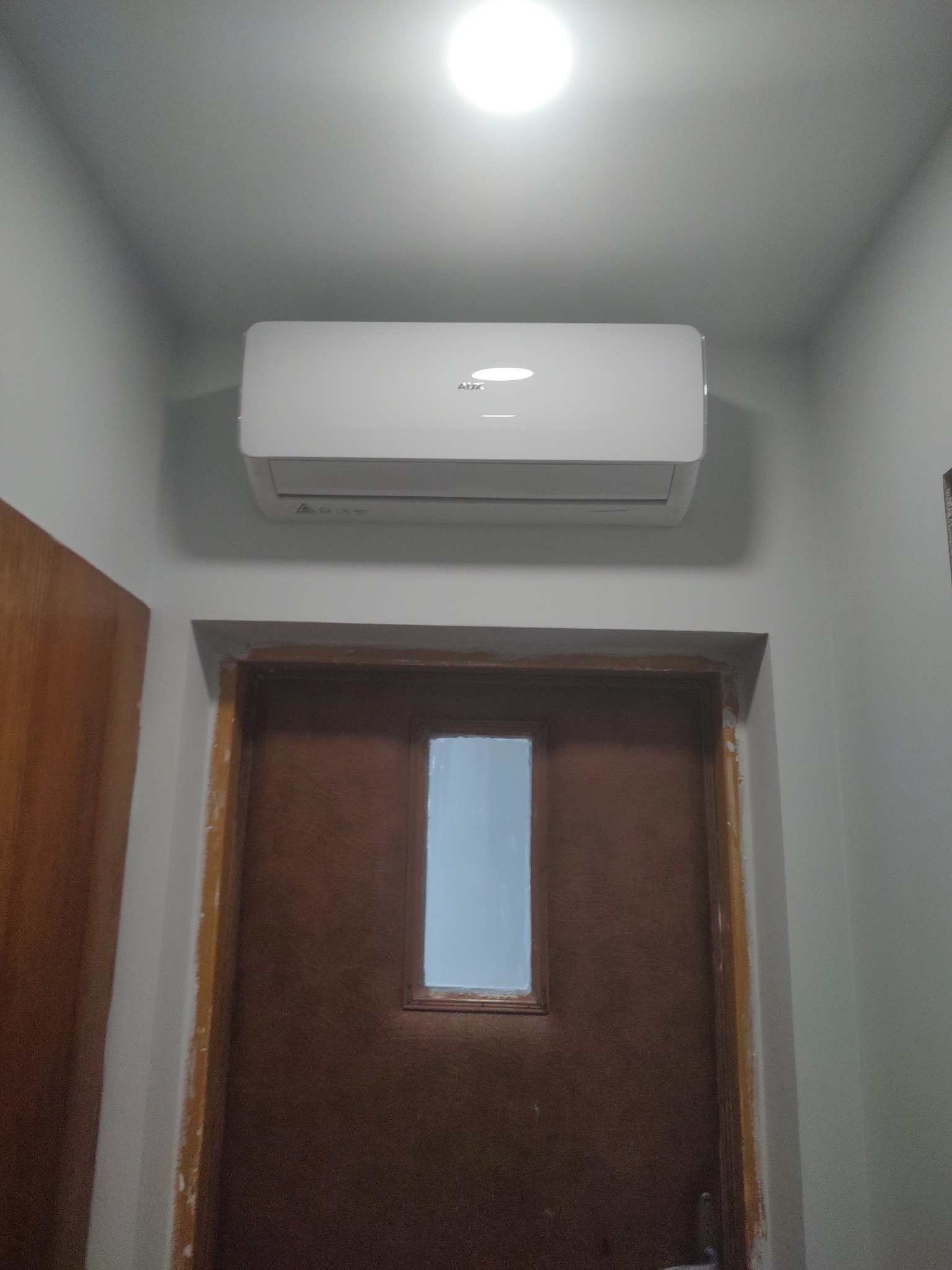 Zamontowany klimatyzator ścienny marki AUX w pomieszczeniu z brązowymi drzwiami i białą ścianą, oświetlony górnym światłem.