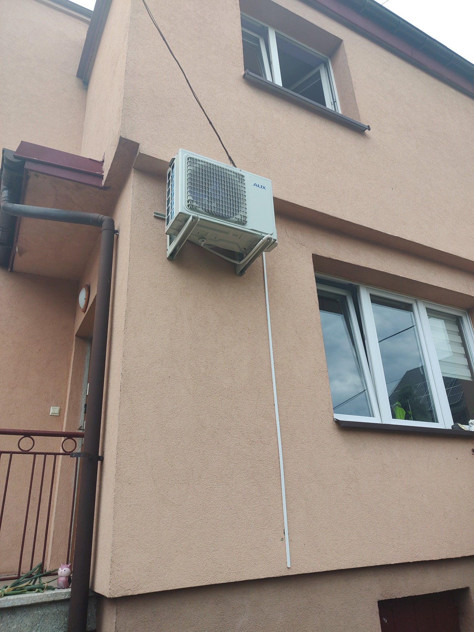 Zamontowana jednostka zewnętrzna klimatyzacji marki AUX na elewacji budynku o beżowej strukturze, widoczny fragment okna i rynny.
