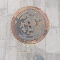 KAMEX Kamil Leśniak - Żeliwna studzienka kanalizacyjna z wzorem labiryntu wkomponowana w szarą kostkę brukową, obok widoczny fragment buta roboczego.