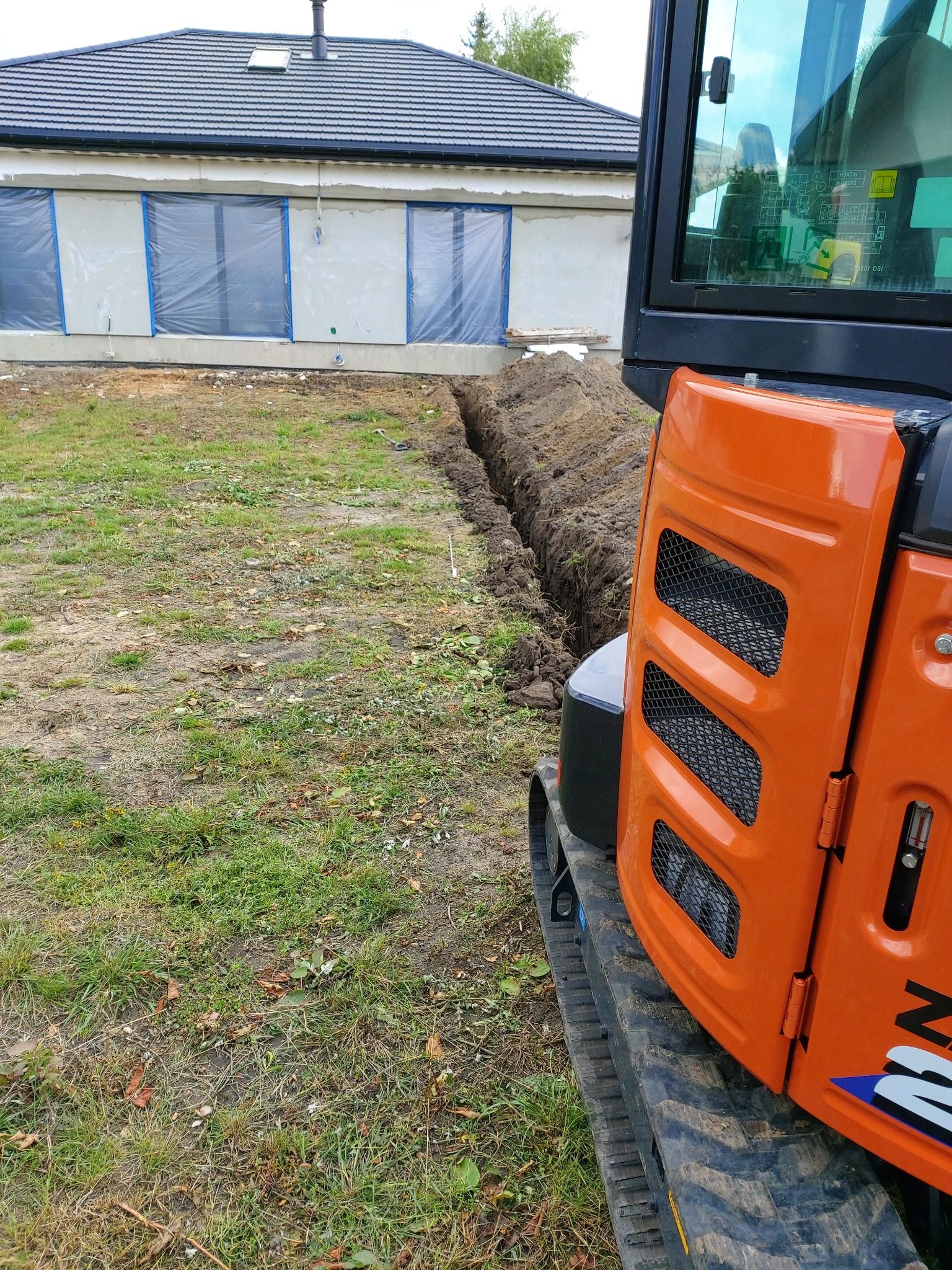 Pomarańczowa minikoparka częściowo widoczna na tle wykopanego wąskiego rowu w trawie, obok budynku w trakcie wykończenia z oknami zabezpieczonymi folią.