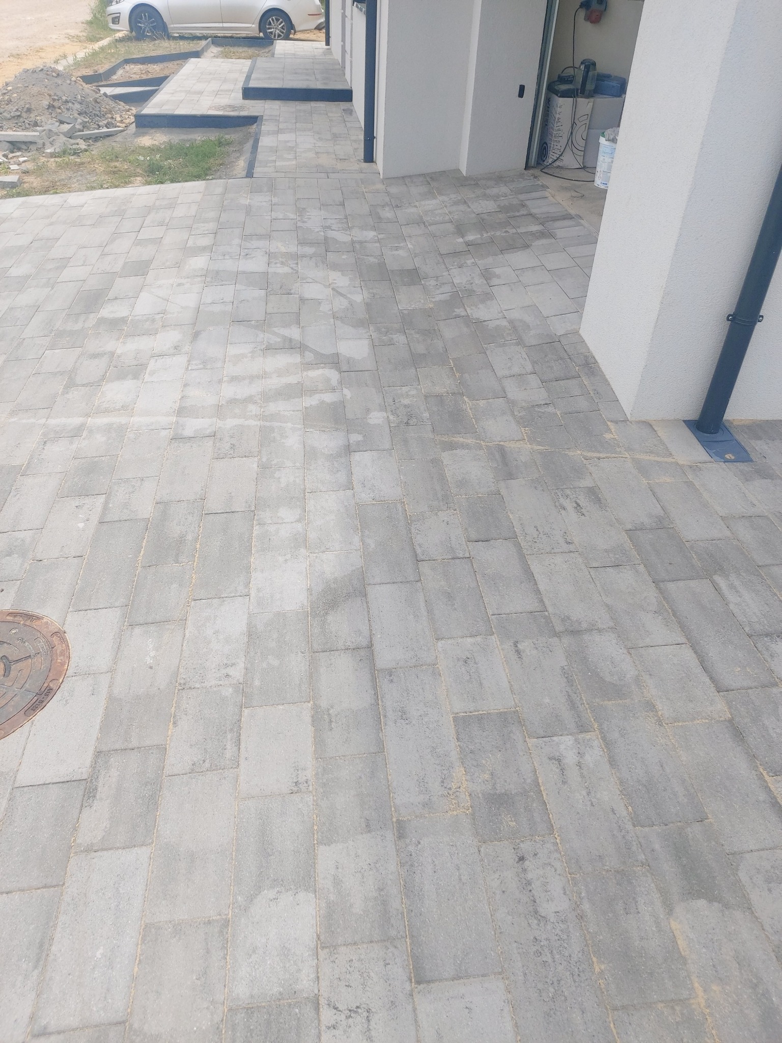 Ułożona szara kostka brukowa przed budynkiem, widoczny fragment samochodu i studzienka kanalizacyjna
