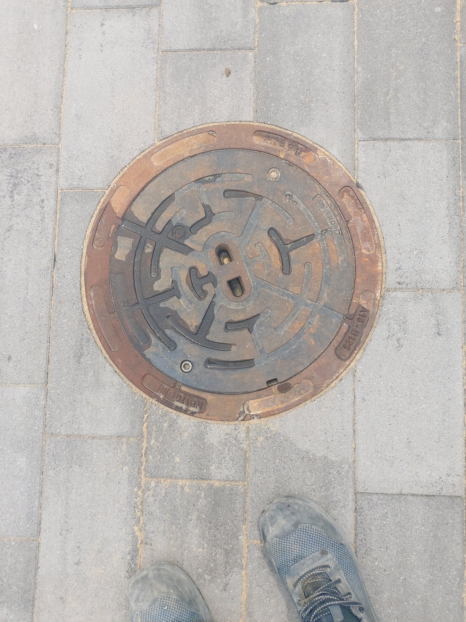 Żeliwna studzienka kanalizacyjna z wzorem labiryntu wkomponowana w szarą kostkę brukową, obok widoczny fragment buta roboczego.