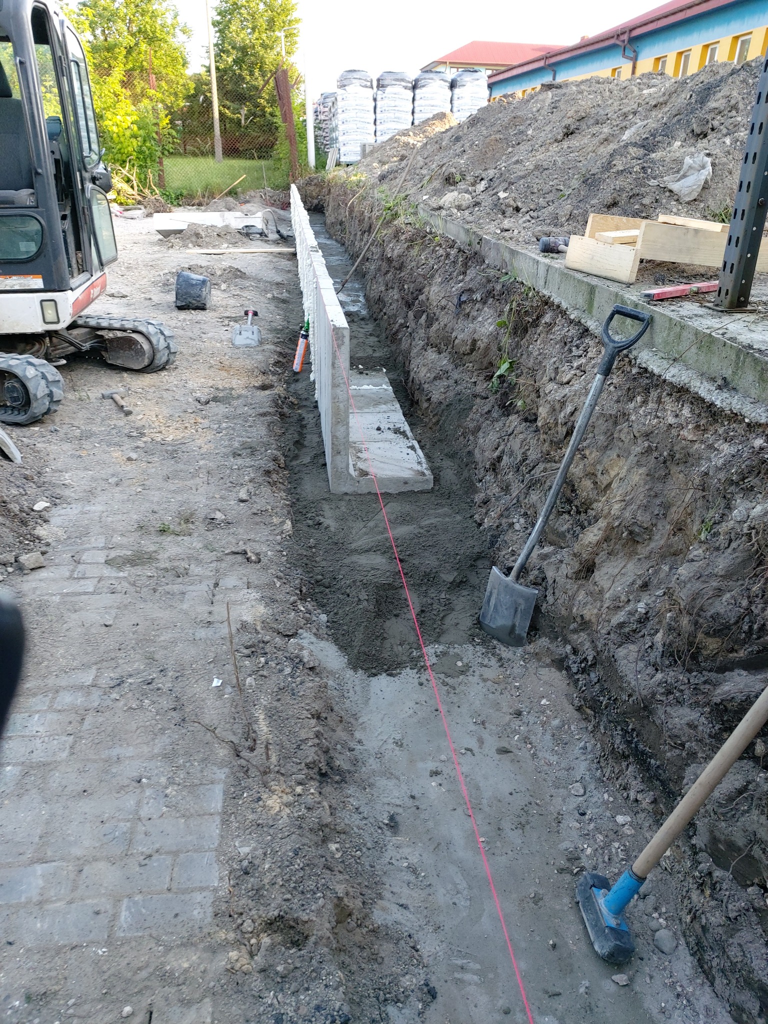 Przygotowanie terenu pod wylewkę fundamentu: wykop w ziemi, szalunek z białych płyt, mini koparka obok, wytyczona linia, stos worków w tle.