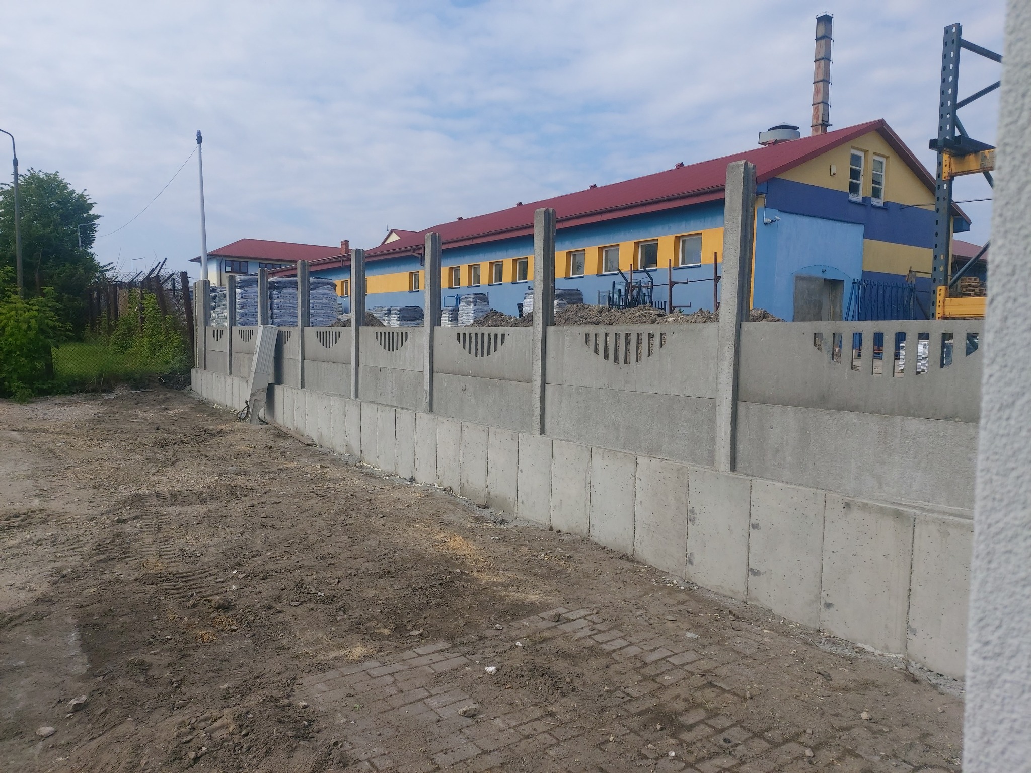 Nowo postawiony, prefabrykowany betonowy płot z ozdobnymi przęsłami, widok na budynek przemysłowy z czerwonym dachem i plac budowy z workami materiałów.