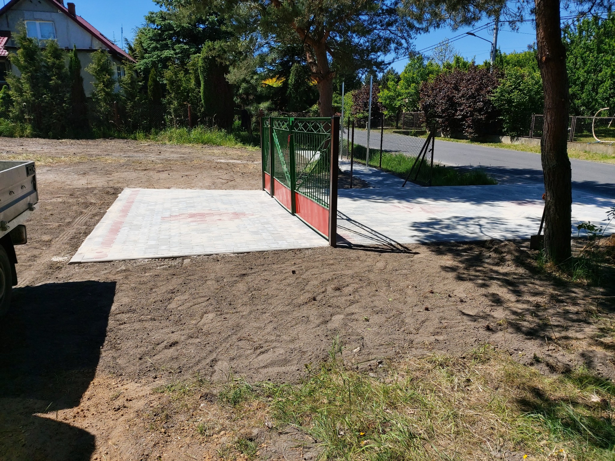 Ułożona nawierzchnia z jasnoszarej kostki brukowej z czerwonym akcentem, prowadząca do zielono-czerwonej bramy wjazdowej, otoczona ziemią i trawą, częściowo zacieniona przez drzewa.