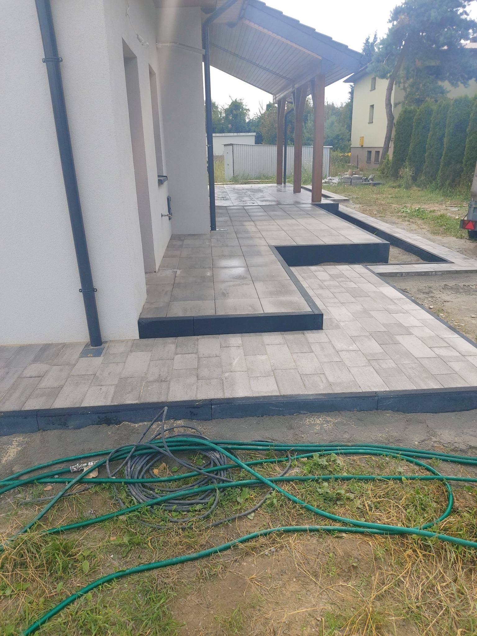 Ułożona szara kostka brukowa z ciemnymi obrzeżami przy ścianie budynku i zadaszeniu, widoczne zwinięte węże ogrodowe na trawie.