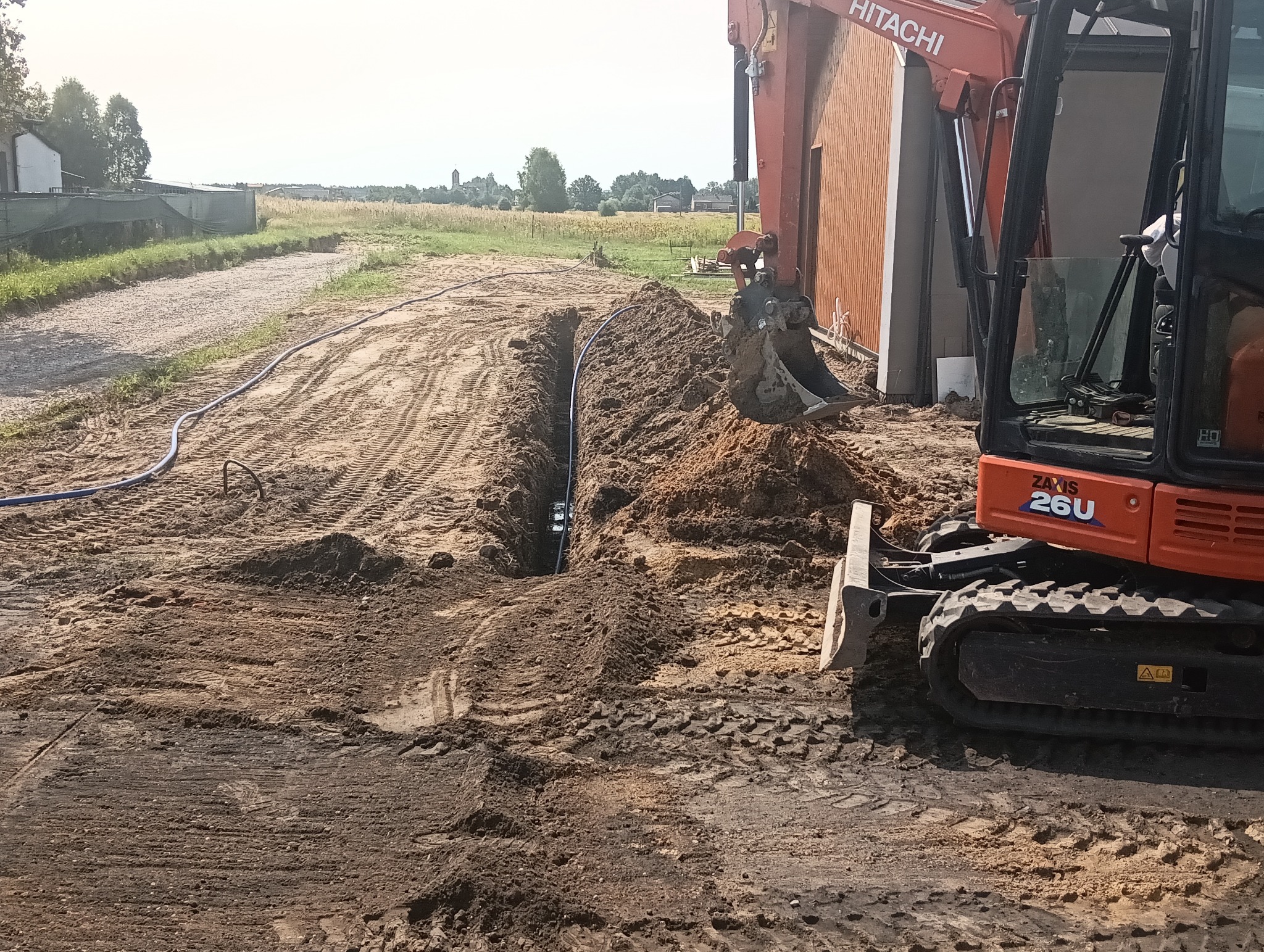 Pomarańczowa koparka Hitachi przy wykopie z rurą, na tle polnego krajobrazu z budynkami i wieżą kościoła w oddali. Ślady opon na ziemi.