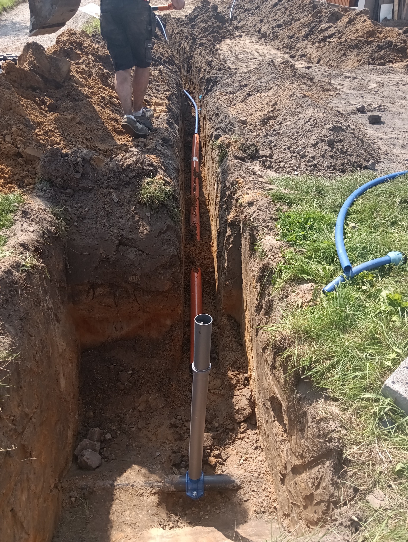 Wykop pod rury wodociągowe w Koniecpolu. Widoczne pomarańczowe i szare rury, niebieski wąż i ziemia. Prace ziemne w toku, widoczny fragment koparki.