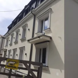 Odnowiona fasada budynku wielorodzinnego z nową stolarką okienną, widoczny fragment rusztowania z żółtą tablicą ostrzegawczą 'Uwaga! Roboty Budowlane'.