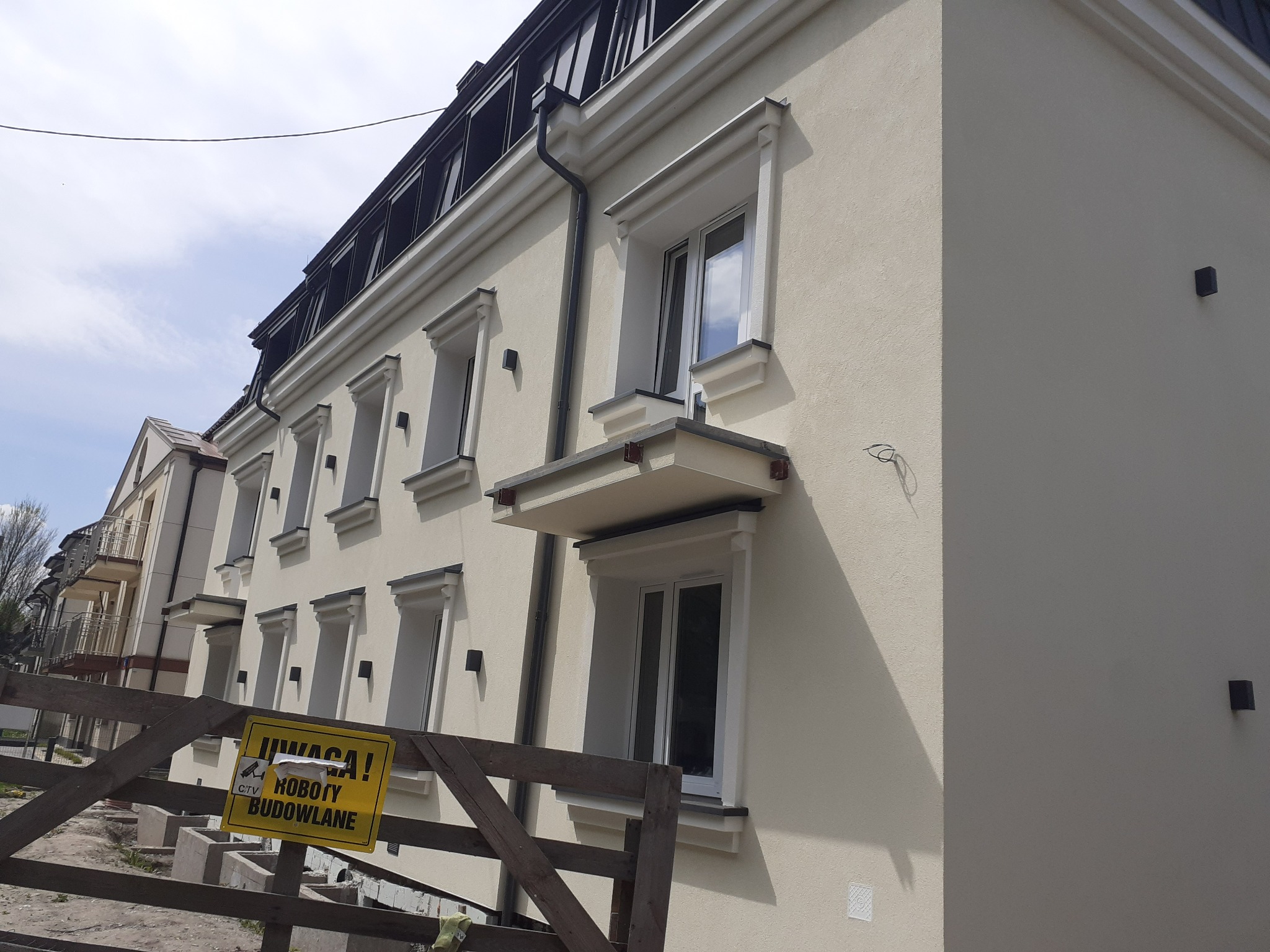 Odnowiona fasada budynku wielorodzinnego z nową stolarką okienną, widoczny fragment rusztowania z żółtą tablicą ostrzegawczą 'Uwaga! Roboty Budowlane'.