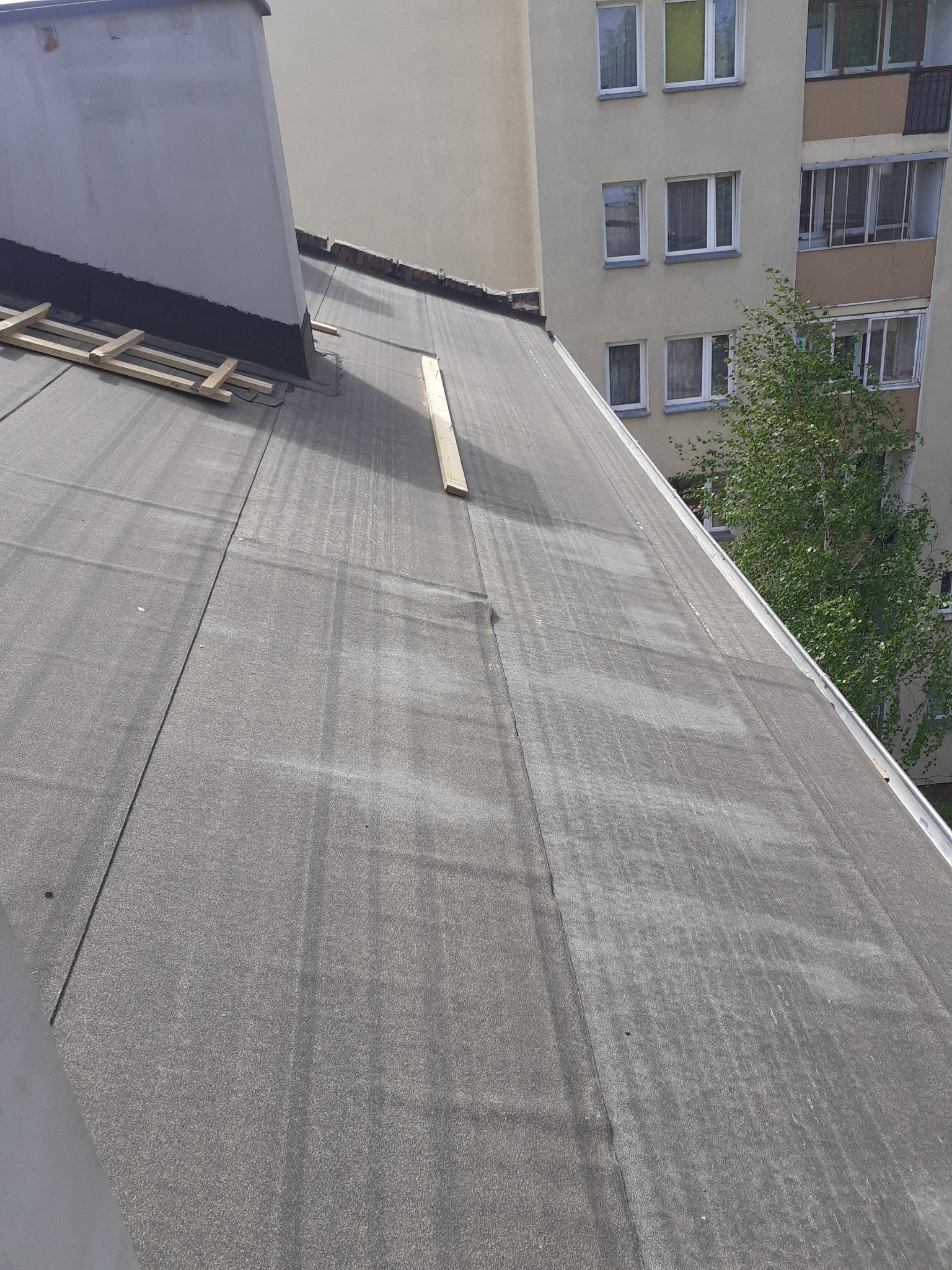 Widok z góry na płaski dach pokryty papą, z drewnianymi łatami przy kominie i fragmentem budynku mieszkalnego w tle.