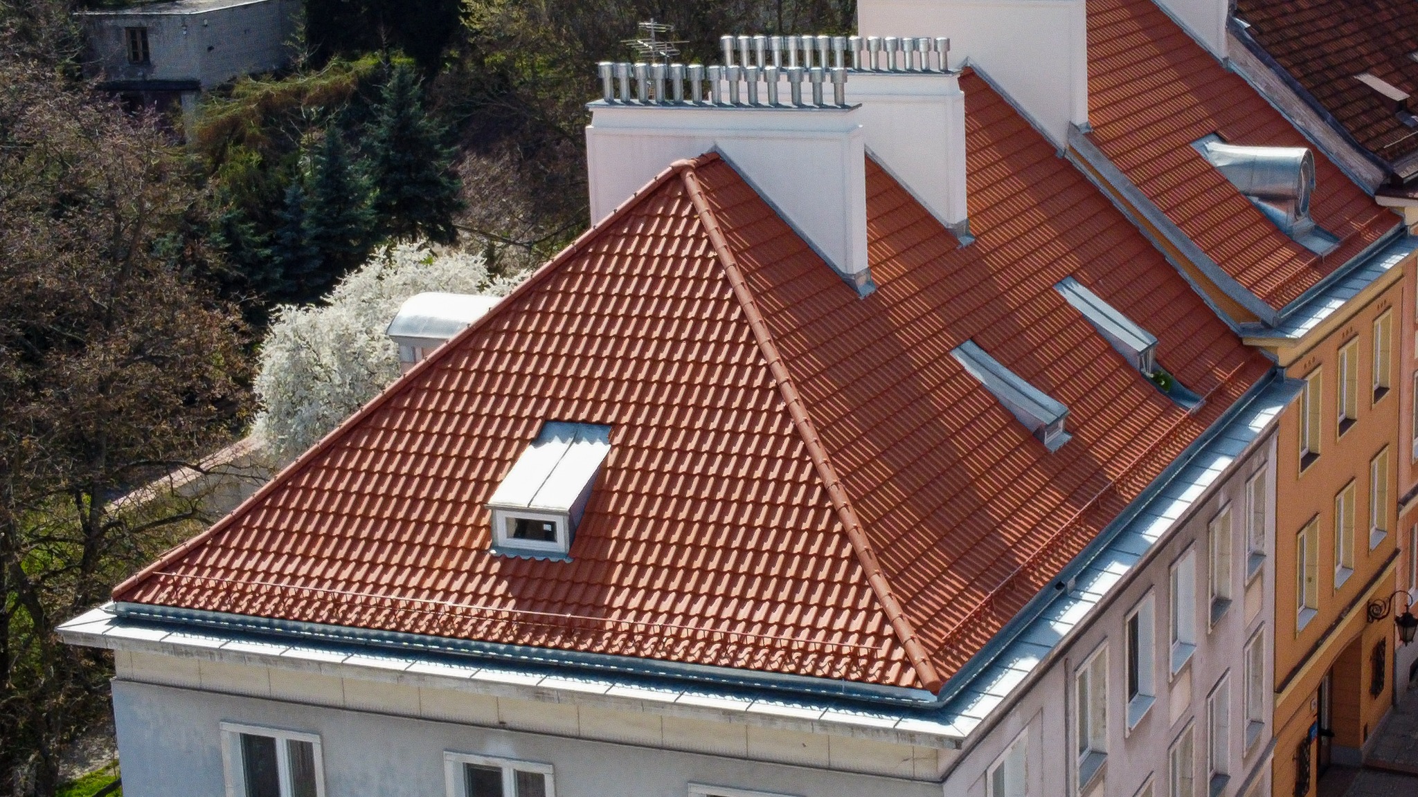 Czerwona dachówka ceramiczna na skośnym dachu budynku mieszkalnego z oknami dachowymi i kominami, widok z góry na tle zieleni i zabudowy miejskiej.