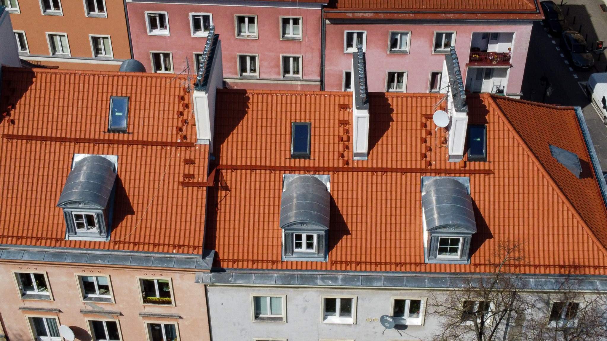 Widok z góry na pomarańczowy dach z dachówką ceramiczną, z lukarnami krytymi blachą w kolorze srebrnym, kominami i oknami dachowymi, na tle kolorowych kamienic.