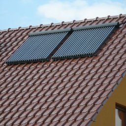 Dariusz Rybacki - Dwa systemy solarne z rurami próżniowymi zamontowane na brązowym dachu z dachówek. Widoczny komin i żółta ściana budynku.
